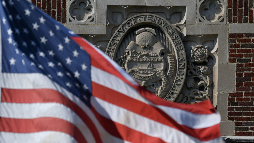 The United States flag flies in the foreground in front of the University seal carved in stone on the front of a UT building