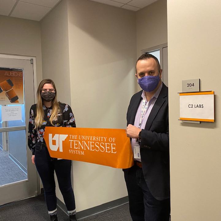 Two IT security professionals in face masks hold an orange ribbon with the UT System logo in front of an office suite
