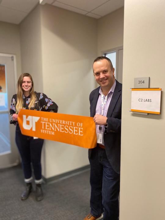Two IT security professionals hold an orange ribbon with the UT System logo in front of an office suite