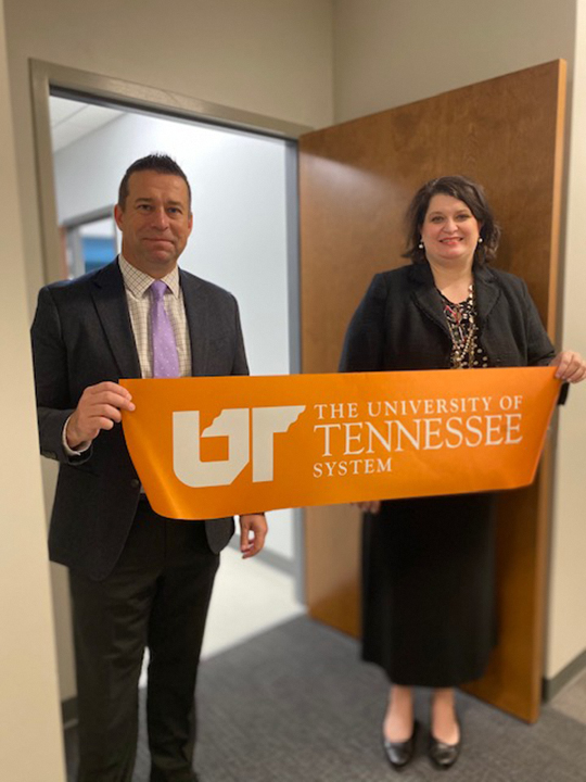 Two doctors in business attire hold up an orange ribbon with the UT System logo