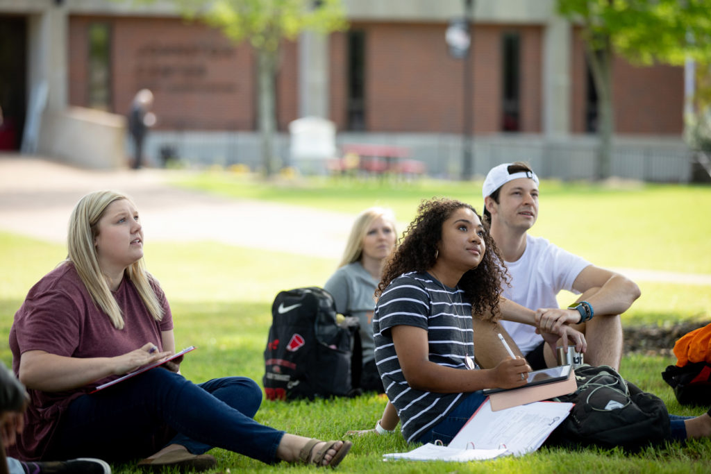 A group of students takes notes and listens to an instructor outdoors in the shade of a tree
